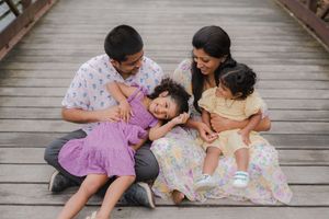 A family of four sits together on a wooden boardwalk, laughing and cuddling during a sunny outdoor photo session.