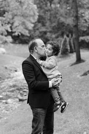 Black and white photo of a man in a blazer kissing a curly-haired toddler on the cheek while holding him near a woodland stream.