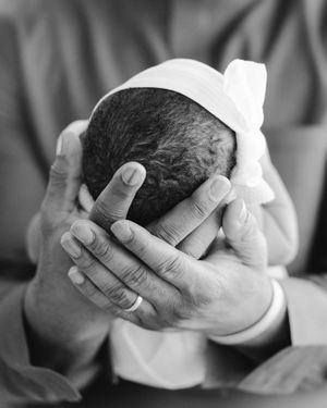 Black and white close-up of a parent's hands gently cradling a newborn baby's head wrapped in a soft knotted headband.