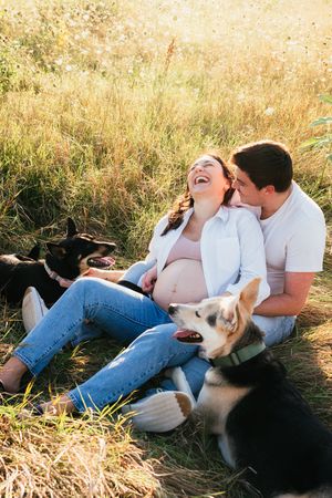 An expecting mother and father sit in a prairie laughing next to their dogs
