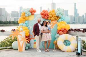 A smiling couple holds their toddler in front of a colorful orange and teal balloon garland with a Chicago skyline backdrop.
