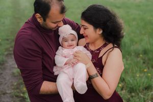 A smiling couple in maroon outfits lovingly hold their baby girl in a pink outfit and bonnet on a grassy field.