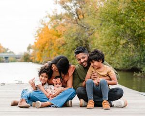 A happy family of five sits close together on a wooden dock by a river with colorful autumn trees in the background.