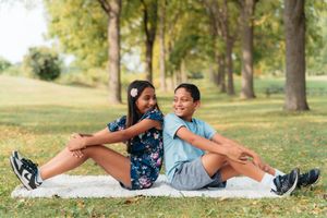 Two smiling children sit back to back on a white blanket in a sunny park surrounded by tall trees and green grass.