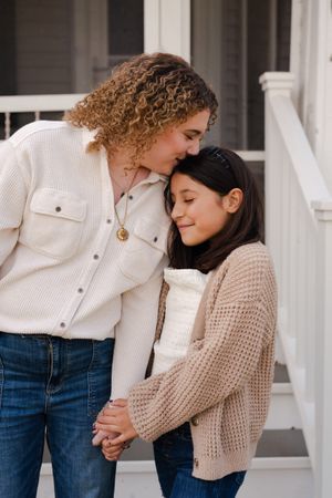 A curly-haired woman gently kisses the forehead of a smiling young girl as they hold hands on white porch steps.