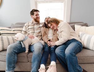 A bearded man and two women laugh together on a gray sofa with a small fluffy white dog in a cozy living room.