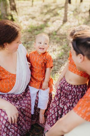 A smiling baby in an orange outfit sits on a woman's lap while two women in matching orange and plum prints share the moment outdoors.