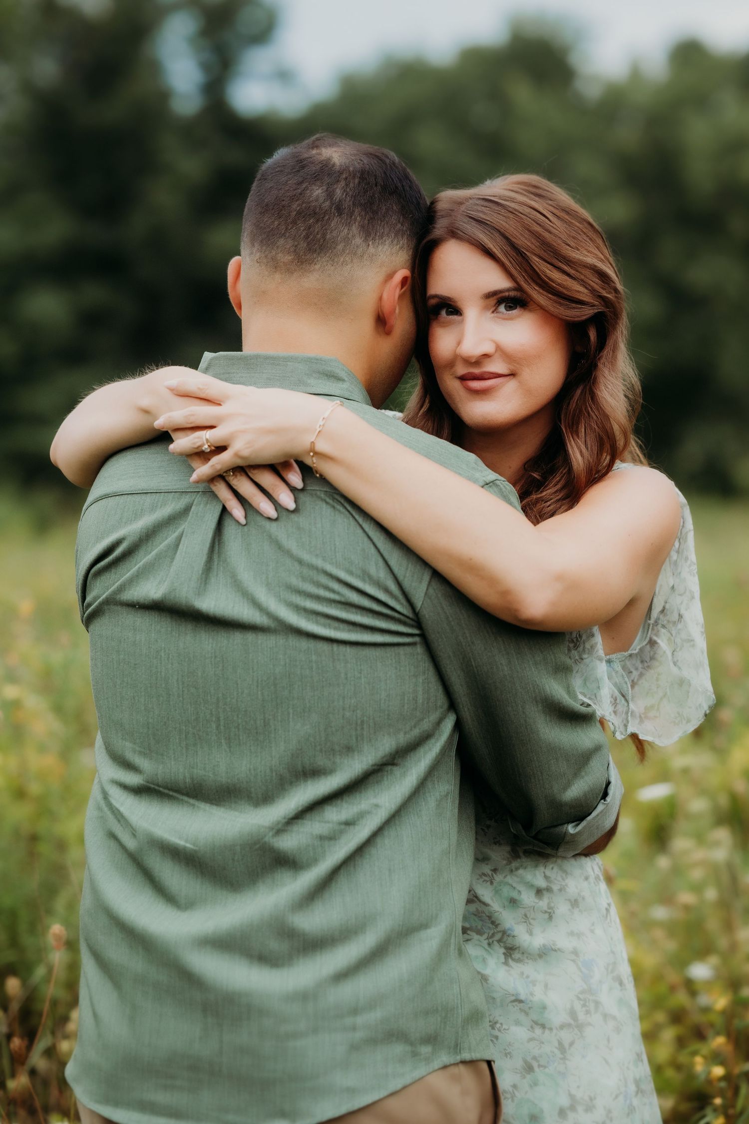 A couple embraces in a wildflower field, woman in floral dress hugging man in green shirt, smiling softly at camera.