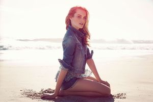 A young woman with red hair sits on a sandy beach wearing a denim jacket, with ocean waves in the background.