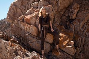 Blonde woman in dark brown outfit sitting on rugged rocky terrain in natural outdoor setting.