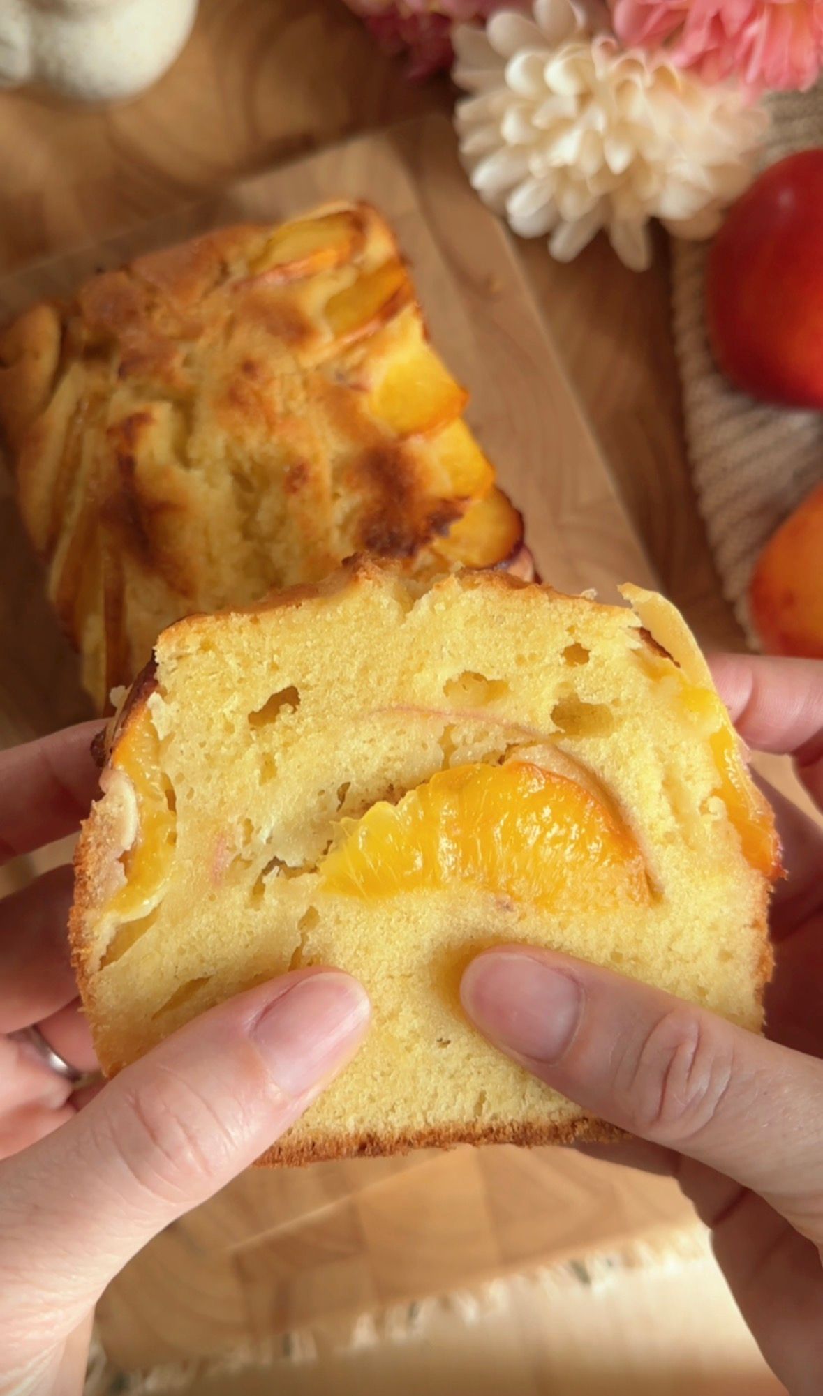 Hands slicing a golden peach pull-apart bread loaf on a wooden board with fresh peaches and pink flowers nearby.