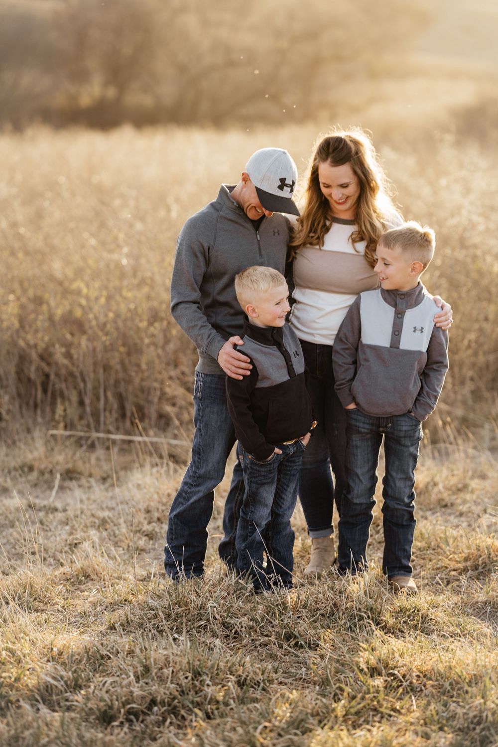 Family of four posing together in a golden field during sunset, parents embracing two young boys in casual outdoor attire.