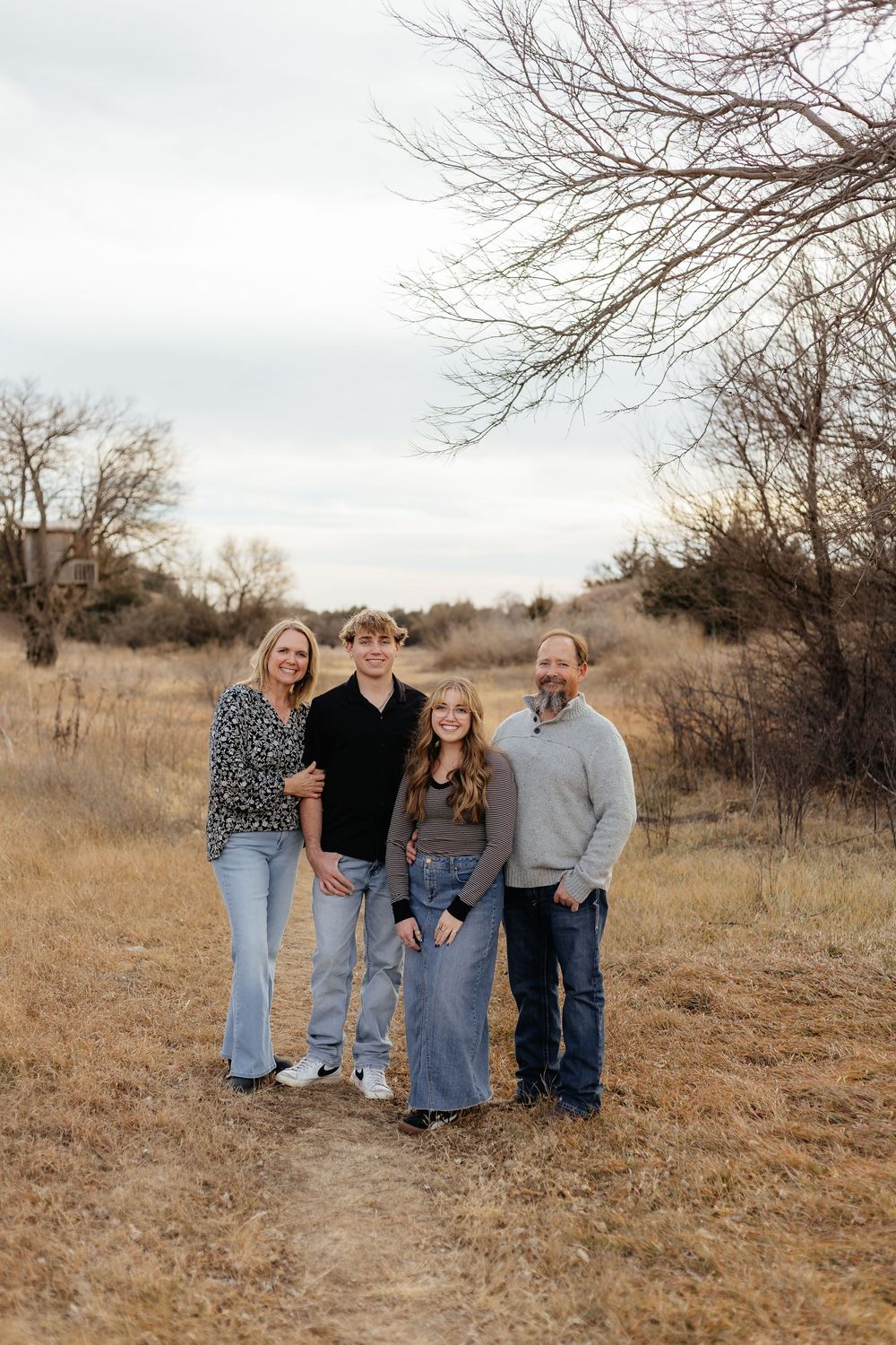 Family of four posing outdoors in a dry winter field with bare trees and overcast sky.