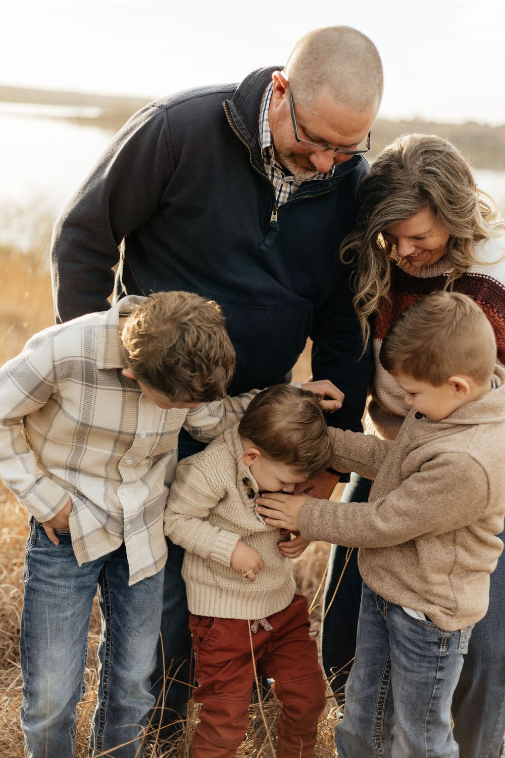 Family of five sharing a joyful moment outdoors in a field, parents and three boys laughing together in cozy fall attire.