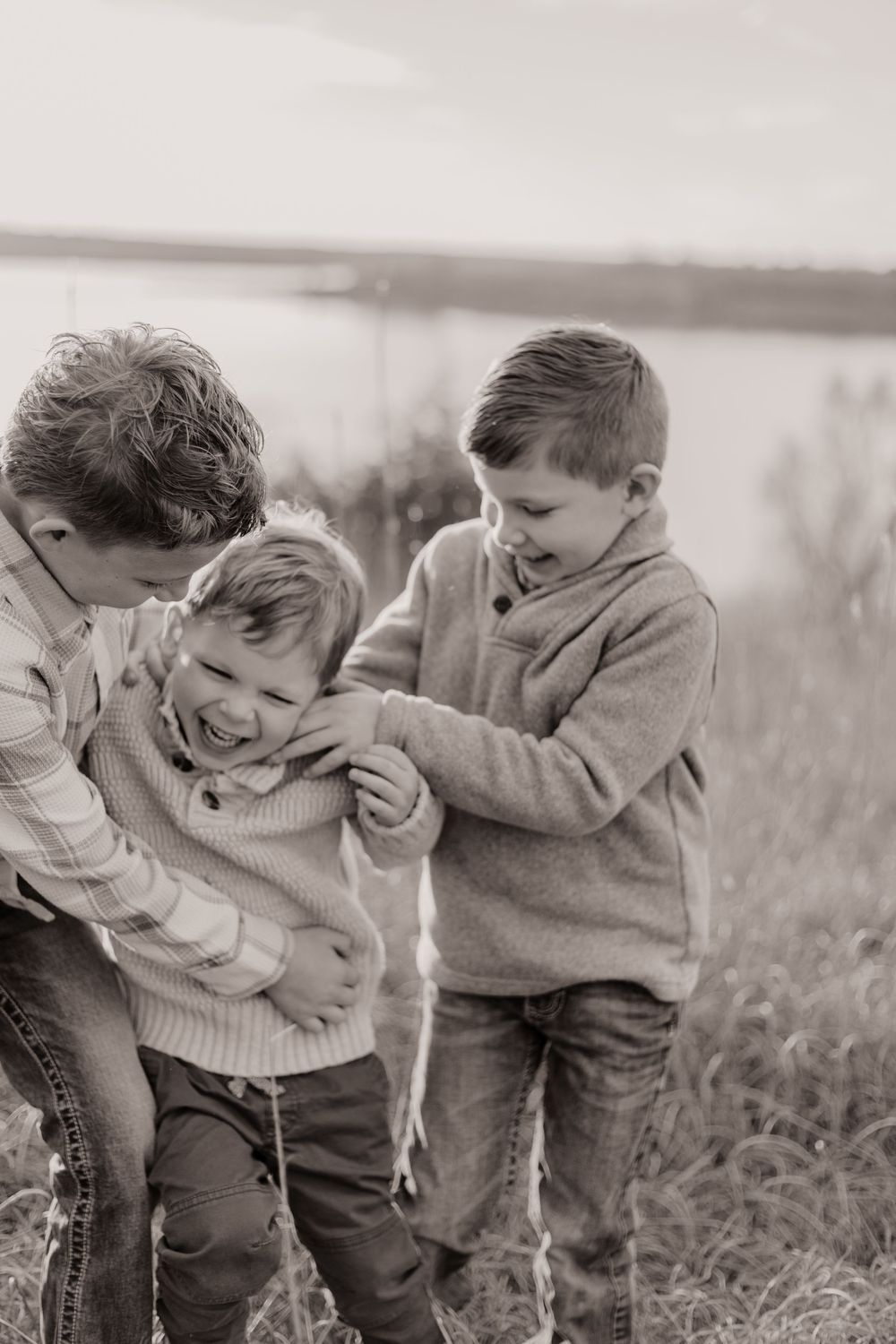Black and white photo of three young boys laughing and playing together outdoors near a lake.