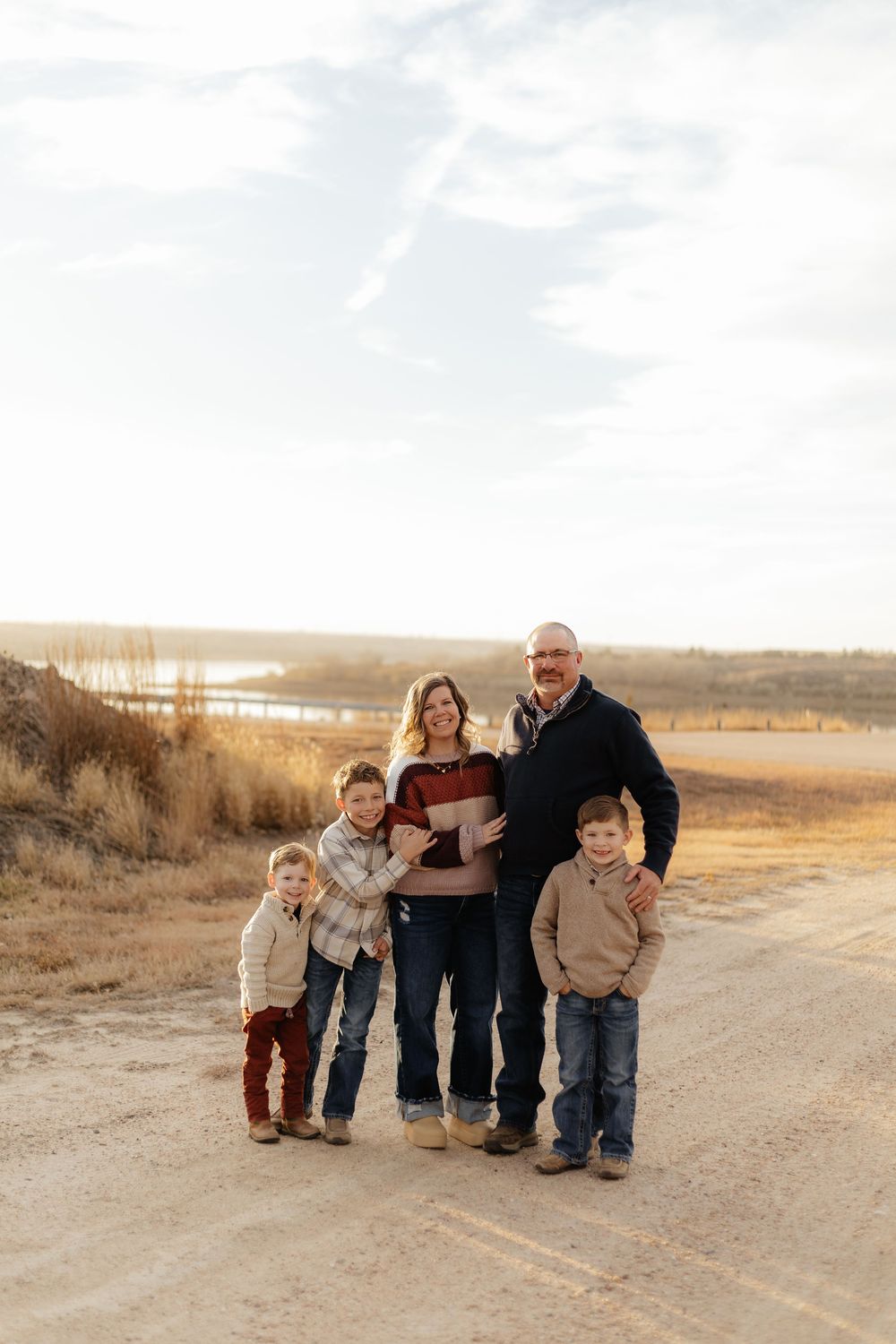 Family of five posing on a rural dirt road during golden hour with a scenic prairie landscape in the background.