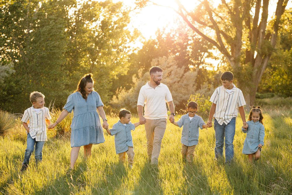 Family of seven holding hands walking through a sunlit meadow at golden hour with tall trees in the background.