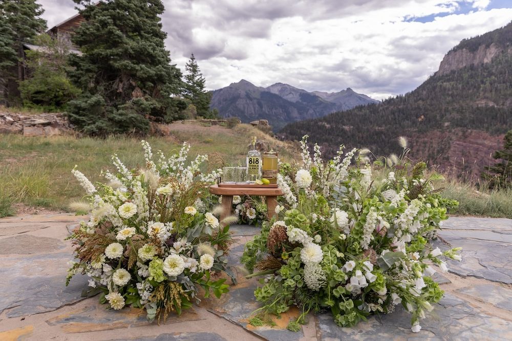 Outdoor wedding altar with white floral arrangements on a stone patio, framed by Rocky Mountain peaks and evergreen trees.