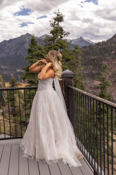 Bride in white tulle gown stands on mountain balcony with scenic Rocky Mountain views and evergreen trees.
