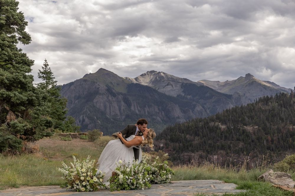 Couple sharing a romantic dip kiss at a mountain wedding with stunning Rocky Mountain peaks and cloudy skies in the background.