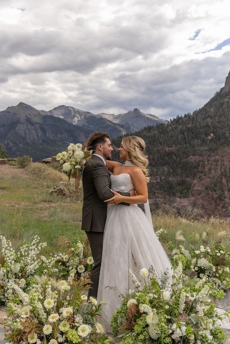 Bride and groom embrace at mountain wedding ceremony with white floral arrangements and dramatic Rocky Mountain backdrop.