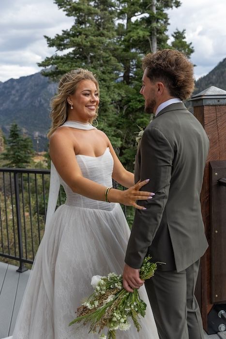 Bride in white strapless gown holding bouquet smiles at groom in gray suit on mountain wedding venue deck.
