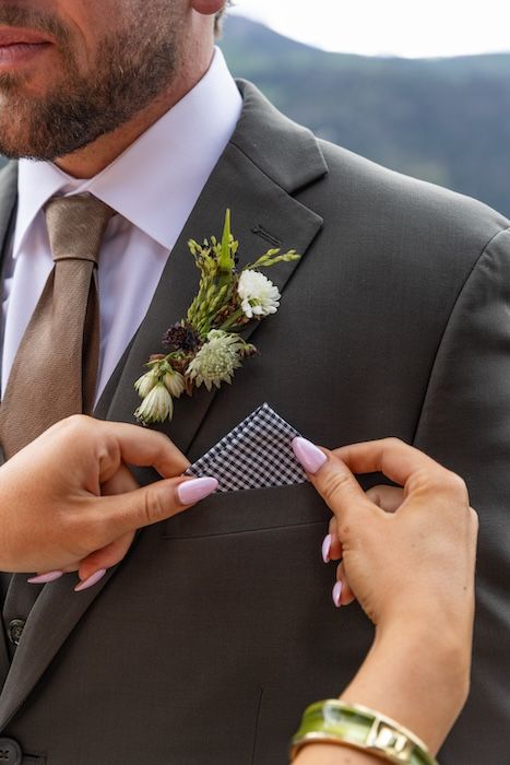 Hands adjusting a gingham pocket square on a groom's gray suit with a floral boutonniere at a wedding.
