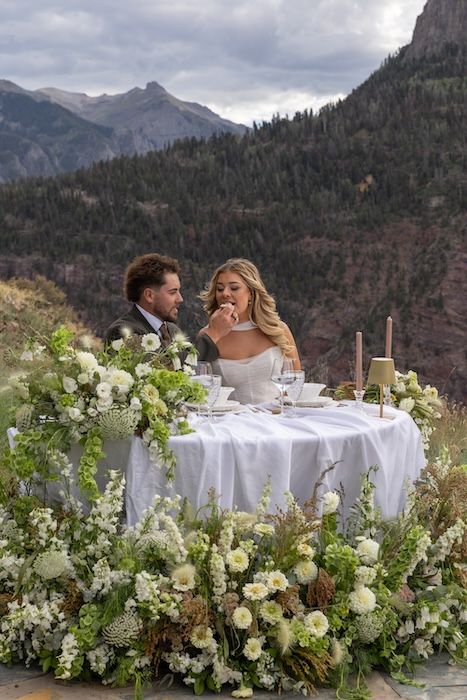Bride and groom share cake at floral-adorned sweetheart table with stunning mountain backdrop at outdoor wedding.