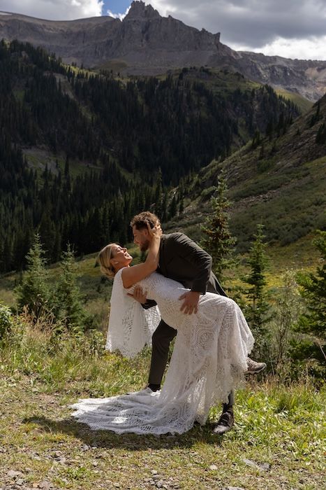 Bride and groom share a romantic dip kiss with stunning mountain and pine forest backdrop during outdoor wedding ceremony.