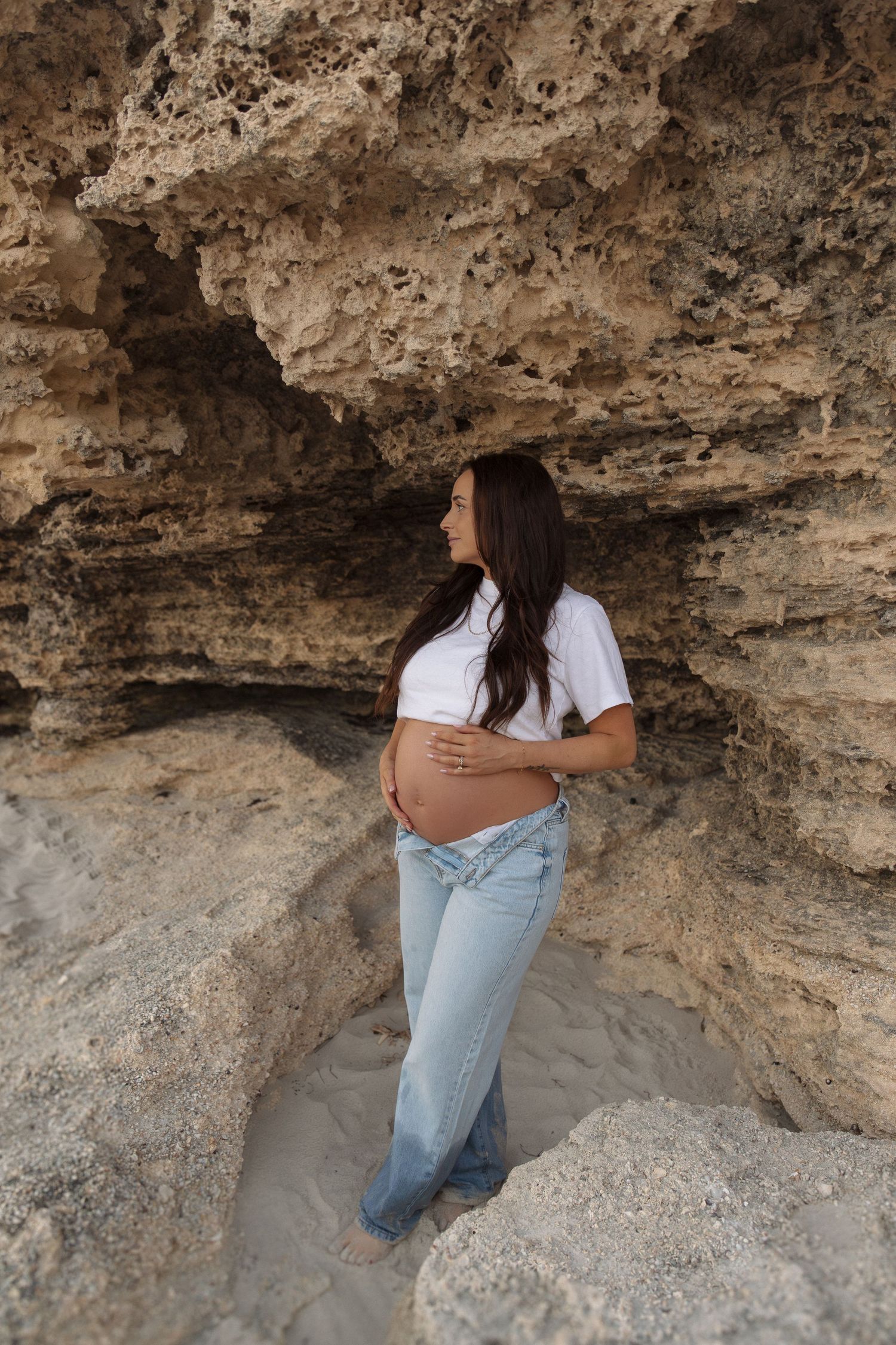 Pregnant woman in white crop top and jeans cradling baby bump against sandy rock formation outdoors.