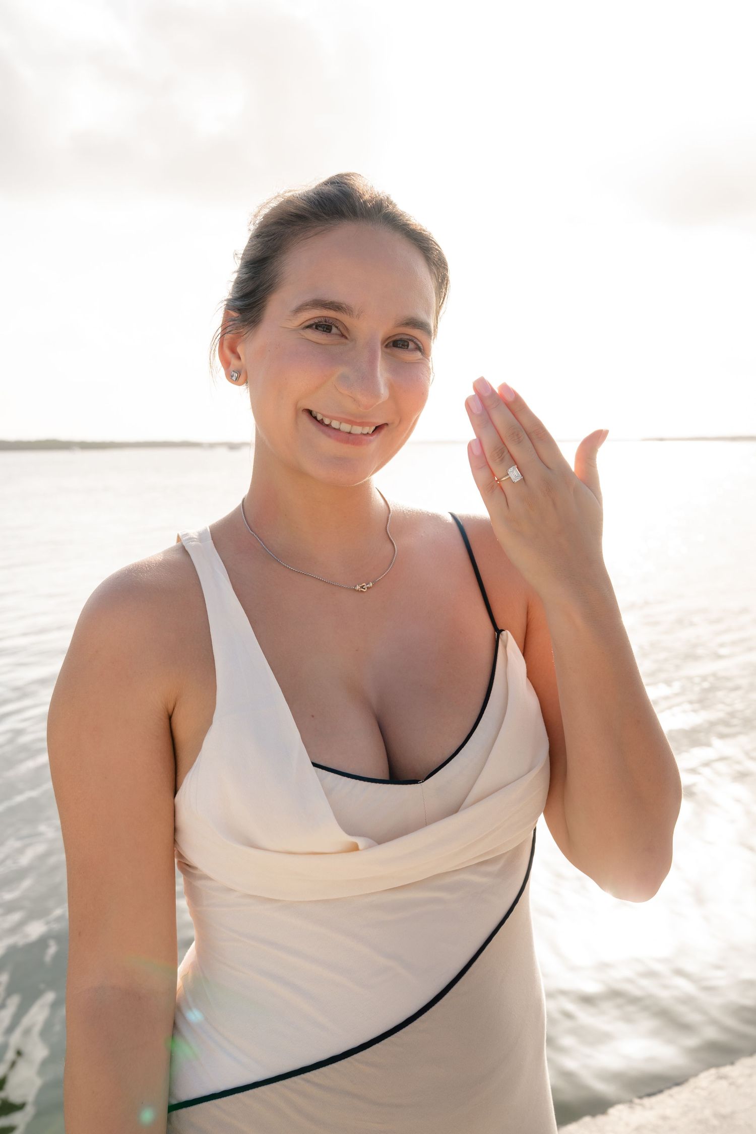Smiling woman in white swimsuit showing ring finger near ocean beach at sunset.