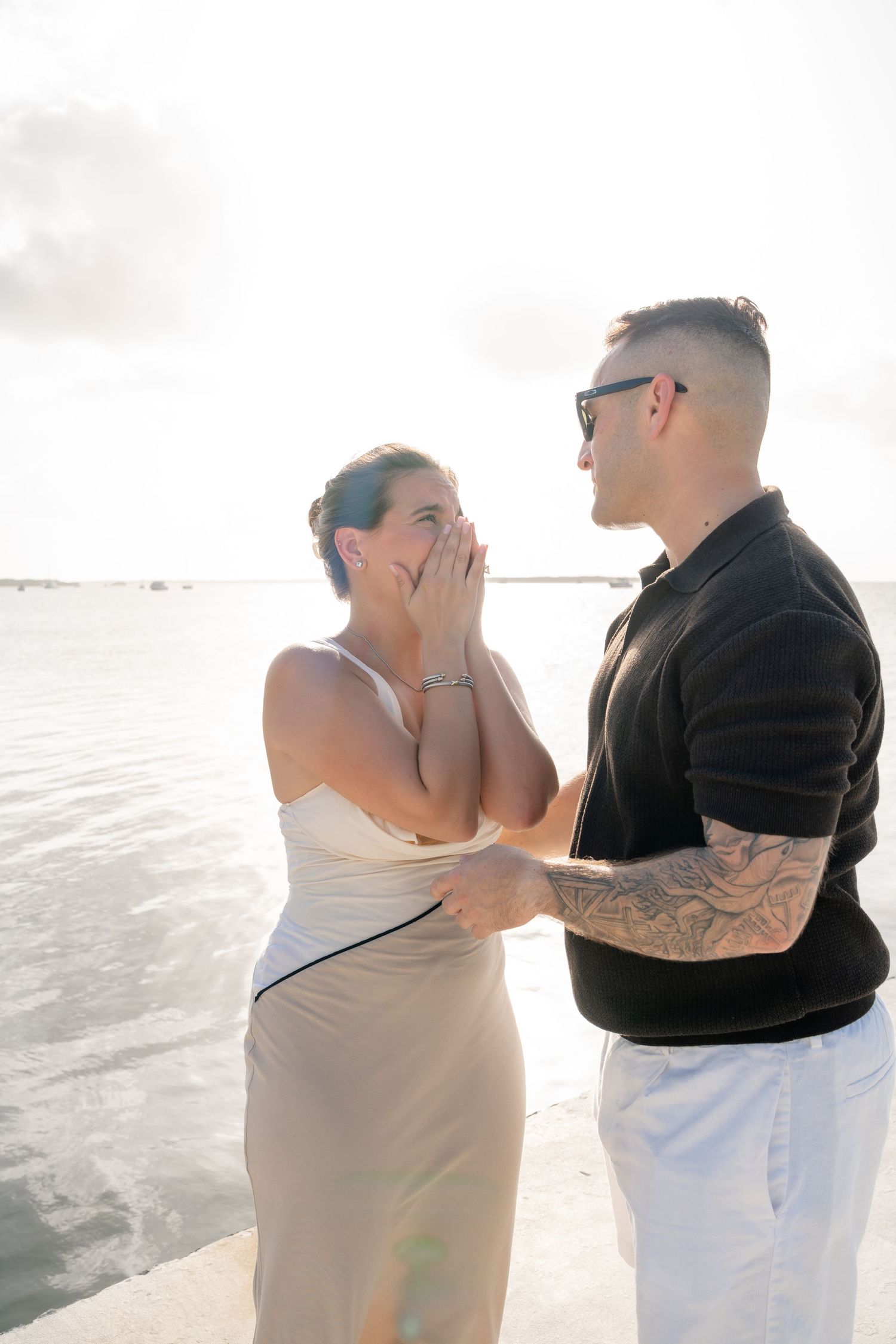 A couple shares an emotional moment by the water, woman in white dress covering her mouth in surprise as tattooed man holds her hands.