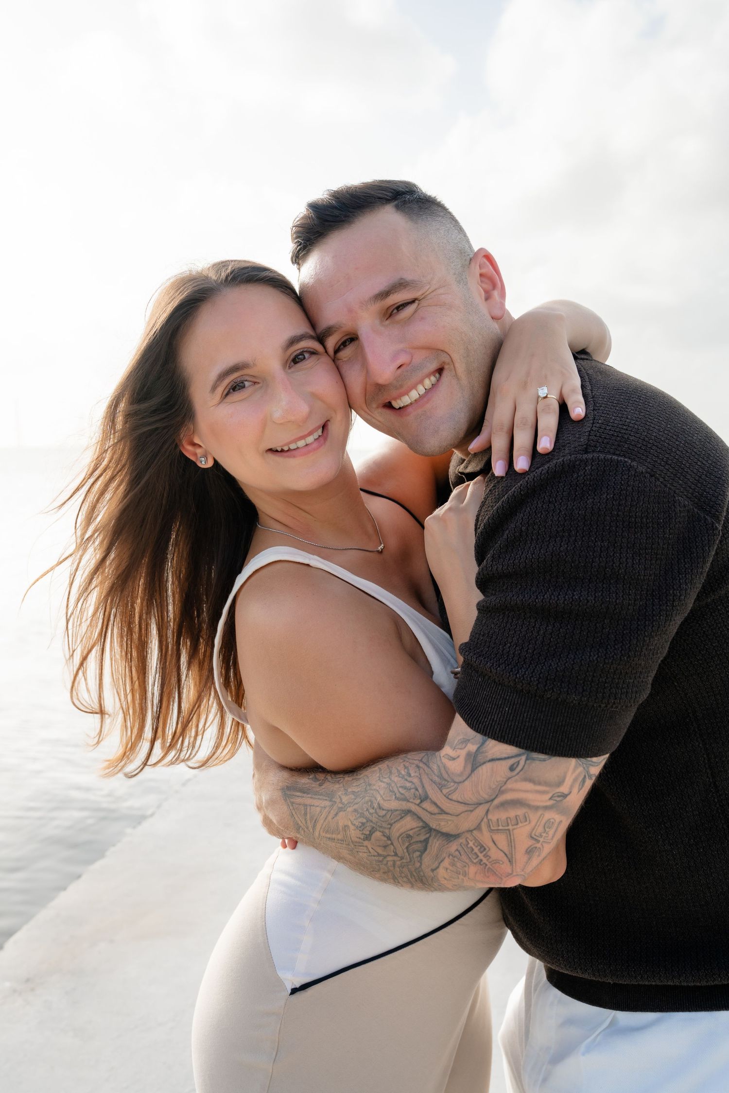 Happy couple embracing on a beach, woman with long hair hugging tattooed man in black sweater, smiling at camera.