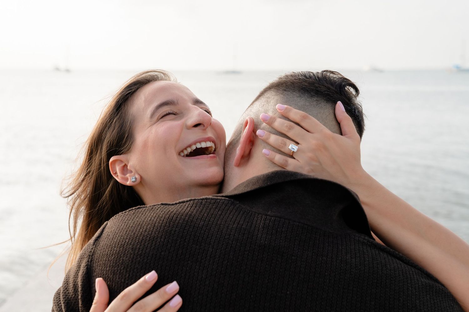 Happy couple embracing on a beach, woman laughing with engagement ring visible, sharing a joyful romantic moment.