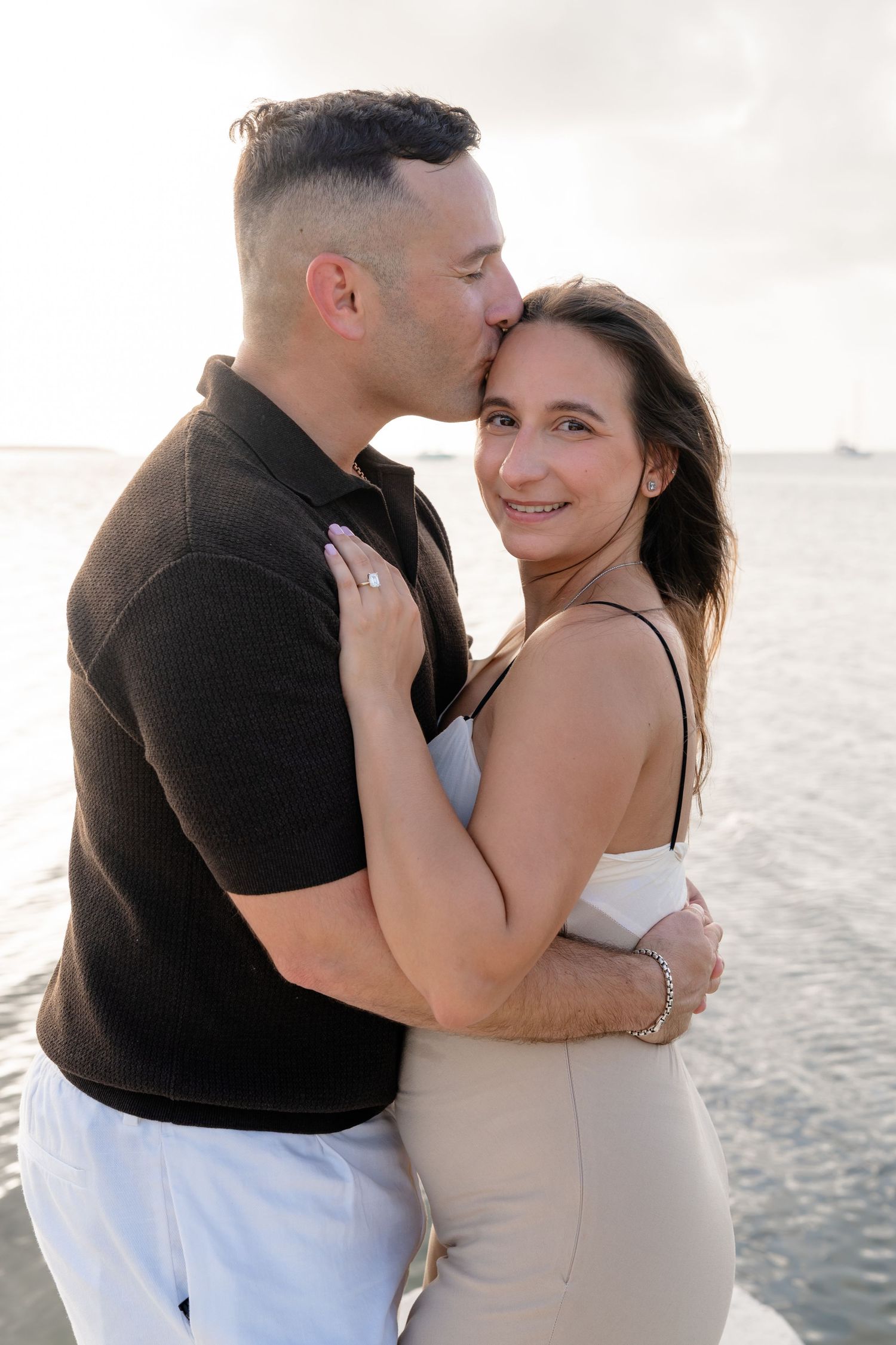 A couple embraces on a beach at sunset, man kissing woman on cheek as she smiles, wearing casual summer attire.