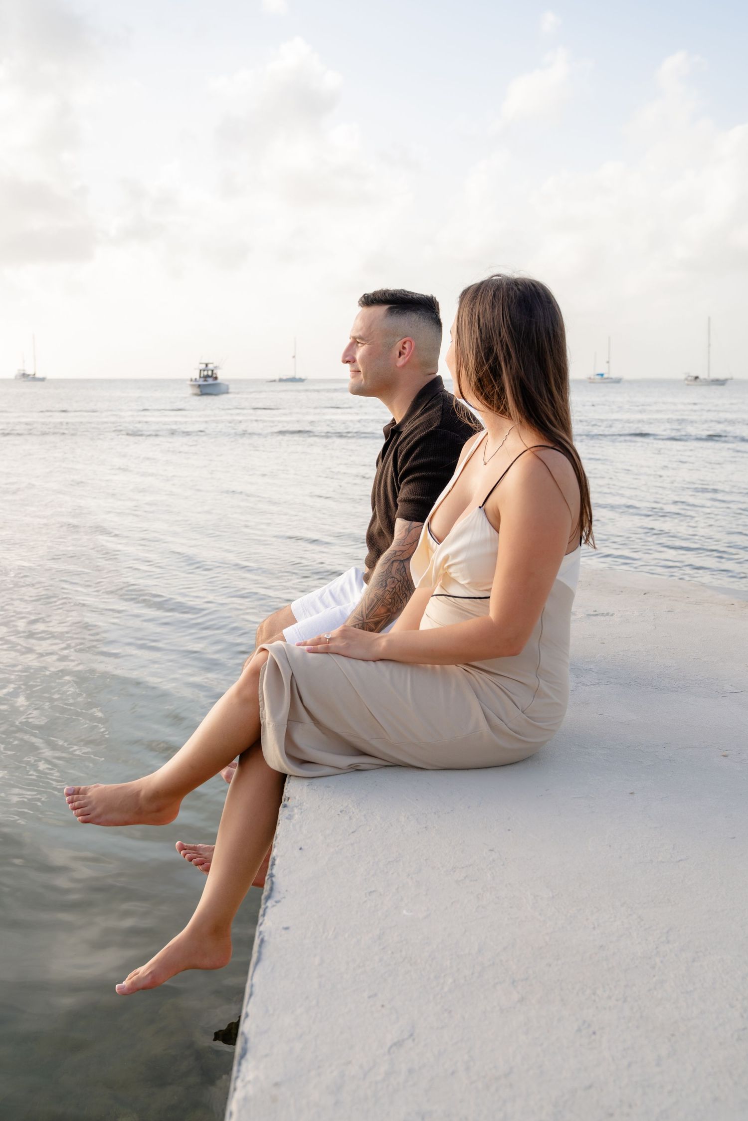 Couple sitting barefoot on a white pier overlooking calm ocean waters with boats in the distance.
