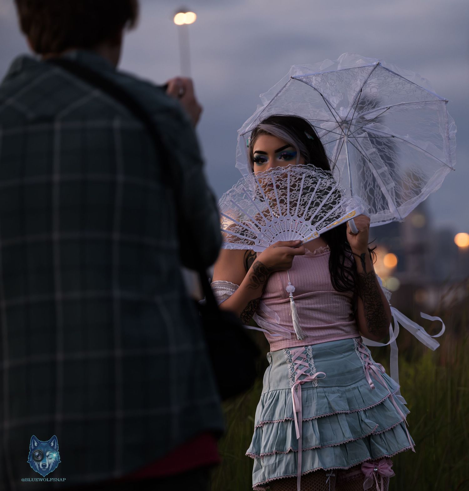 Woman in pink corset and ruffled skirt holds lace fan and parasol outdoors at dusk.