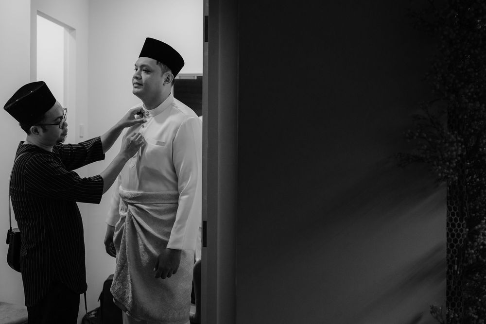 Black and white photo of two men in traditional Malay attire, one adjusting the other's collar indoors.