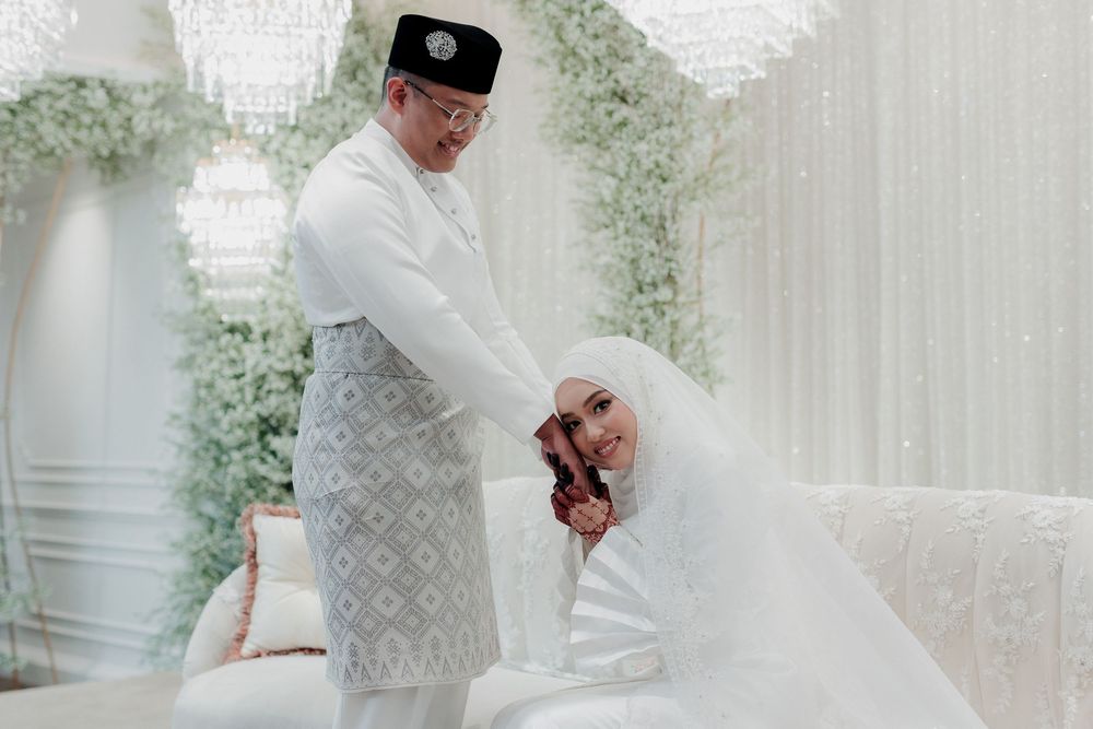 Muslim couple in white traditional attire during a wedding ceremony, groom in songkok cap, bride in hijab and gown.