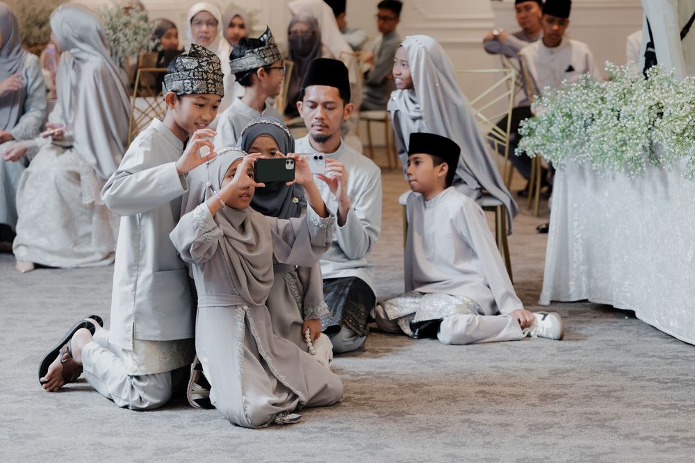 Group of people in traditional Malay attire kneeling during a wedding ceremony, with one person capturing the moment on a phone.