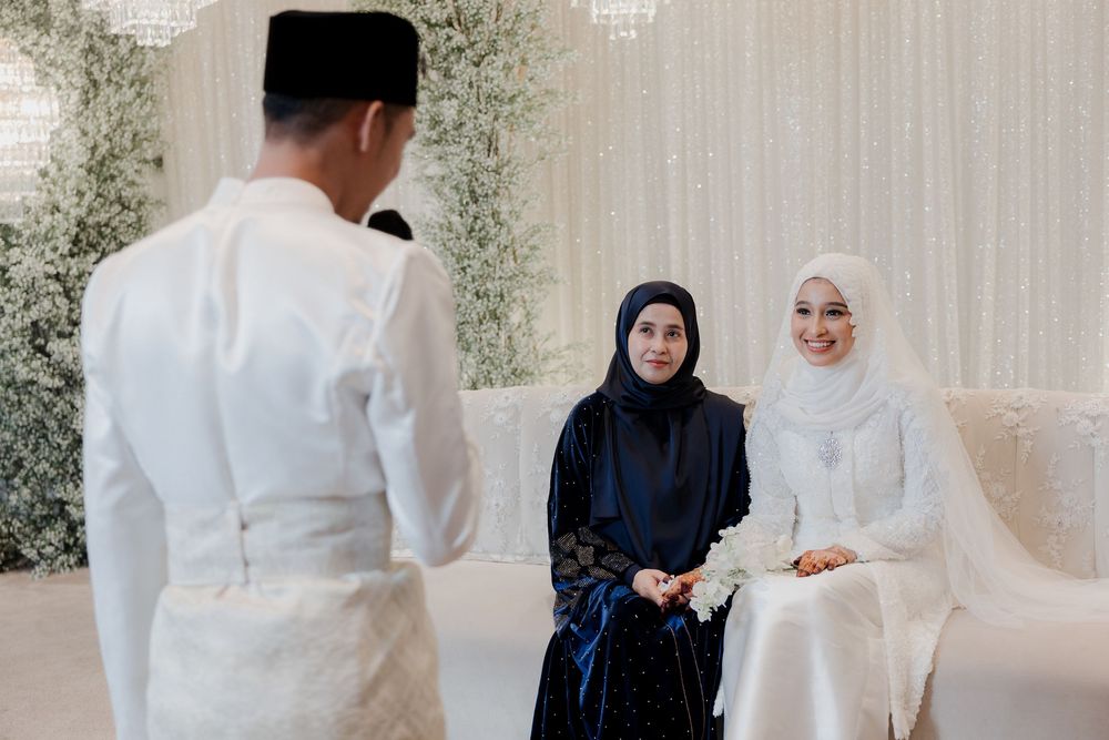 Muslim wedding ceremony with groom in white baju melayu facing two women in hijab, elegant floral backdrop.