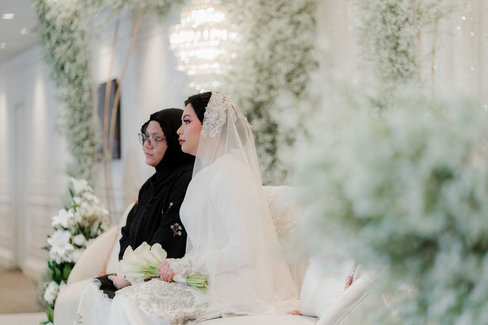 Muslim bride in white hijab and gown sits beside woman in black hijab at elegant floral wedding ceremony.