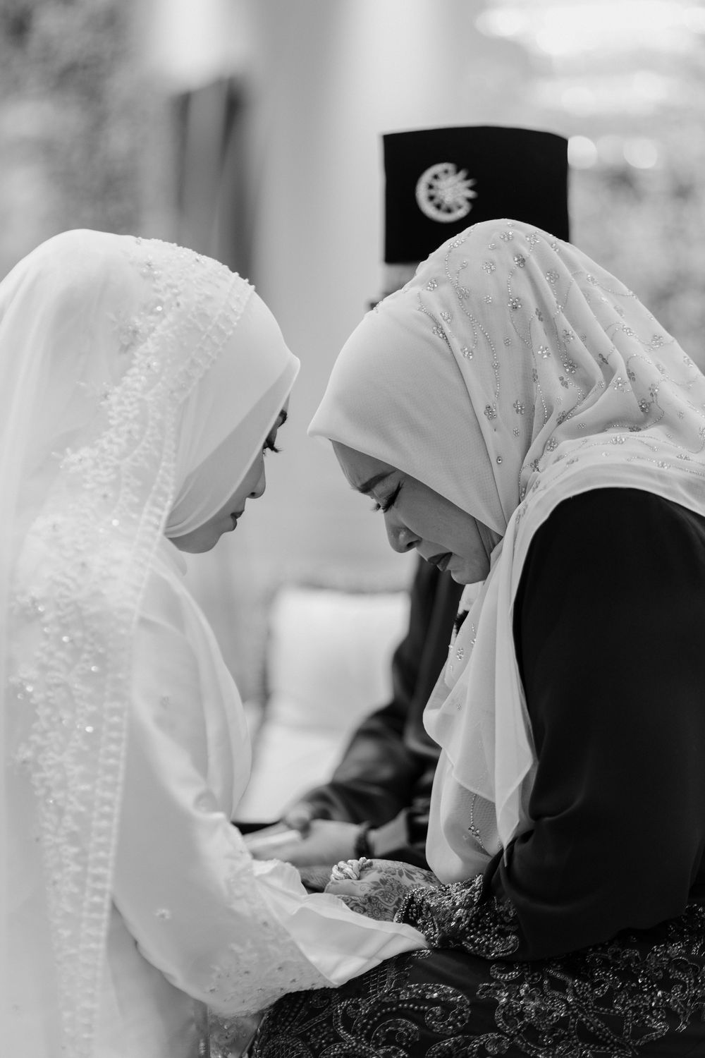Two Muslim women in hijabs share an intimate moment at a wedding ceremony in black and white photography.