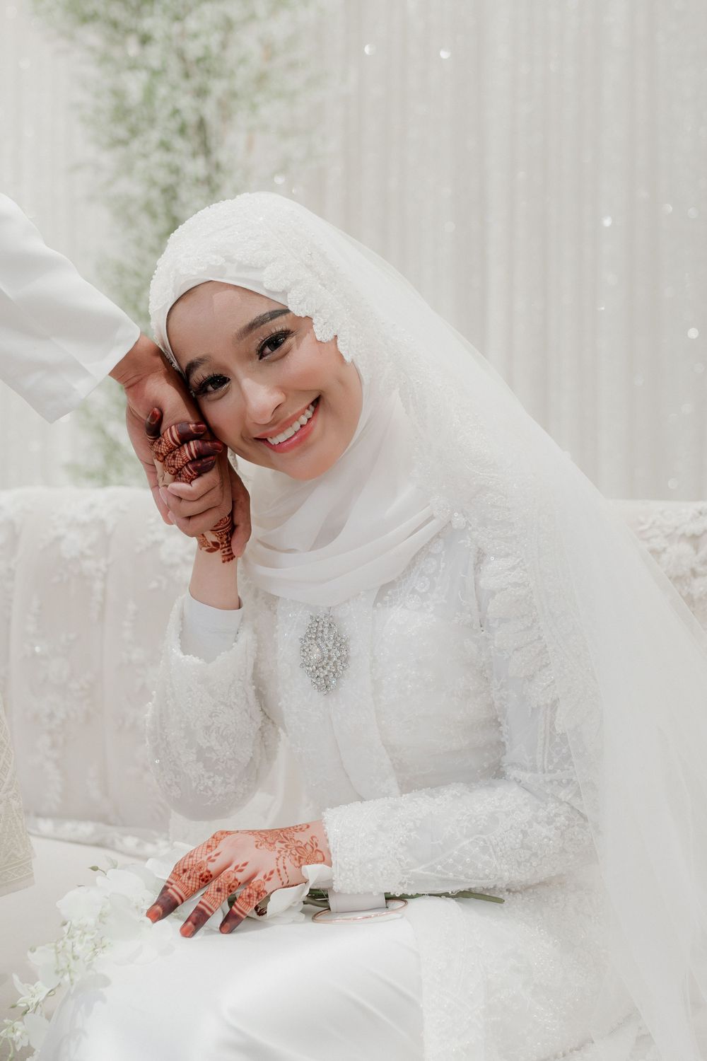 Smiling Muslim bride in white hijab and lace gown with henna hands on wedding day.