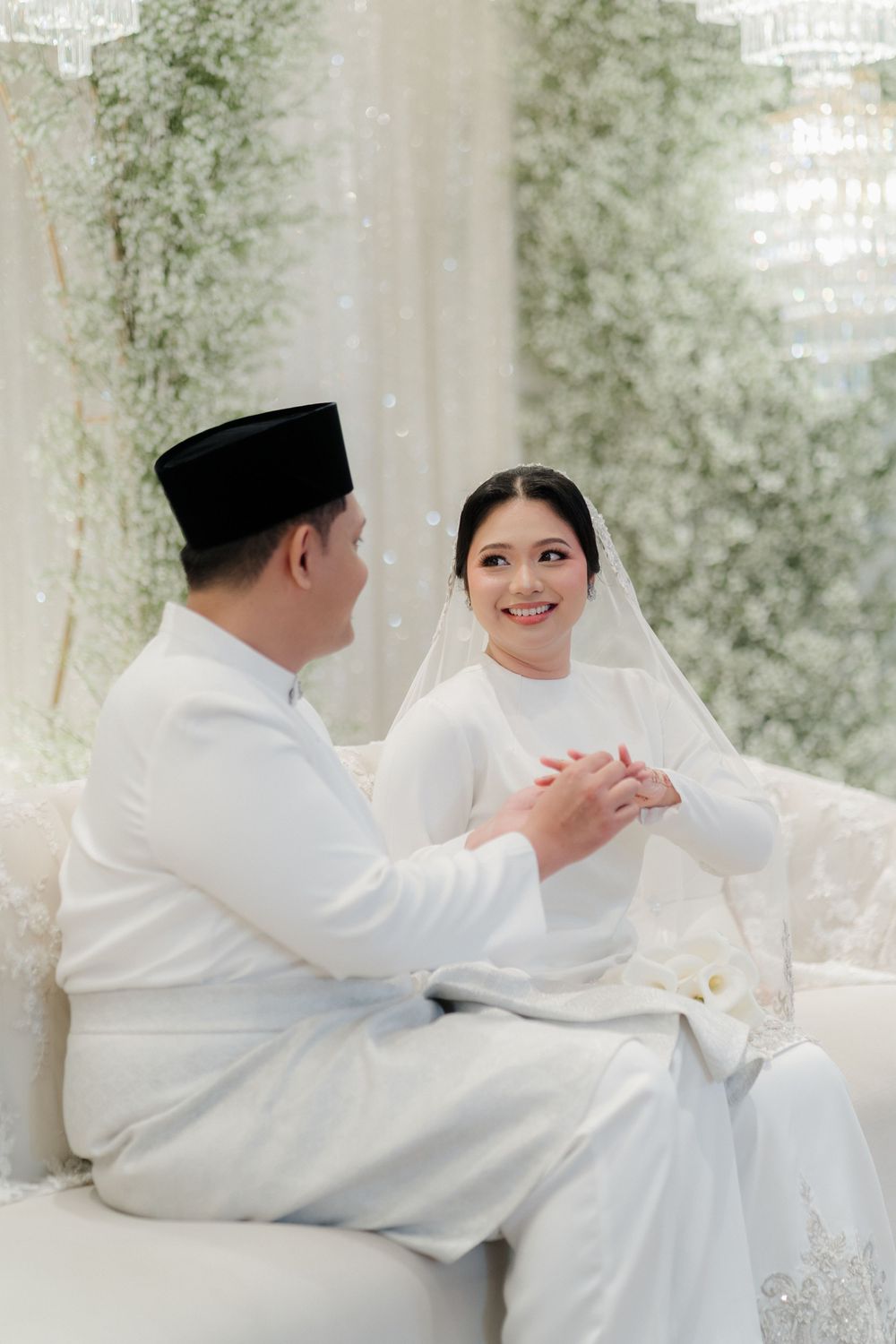 A couple in traditional white Malay wedding attire sit together, holding hands and smiling at each other.