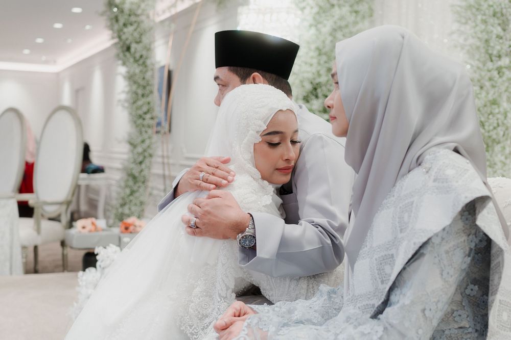 A bride in a white hijab and gown embraces a woman in a gray hijab at an elegant Islamic wedding ceremony.