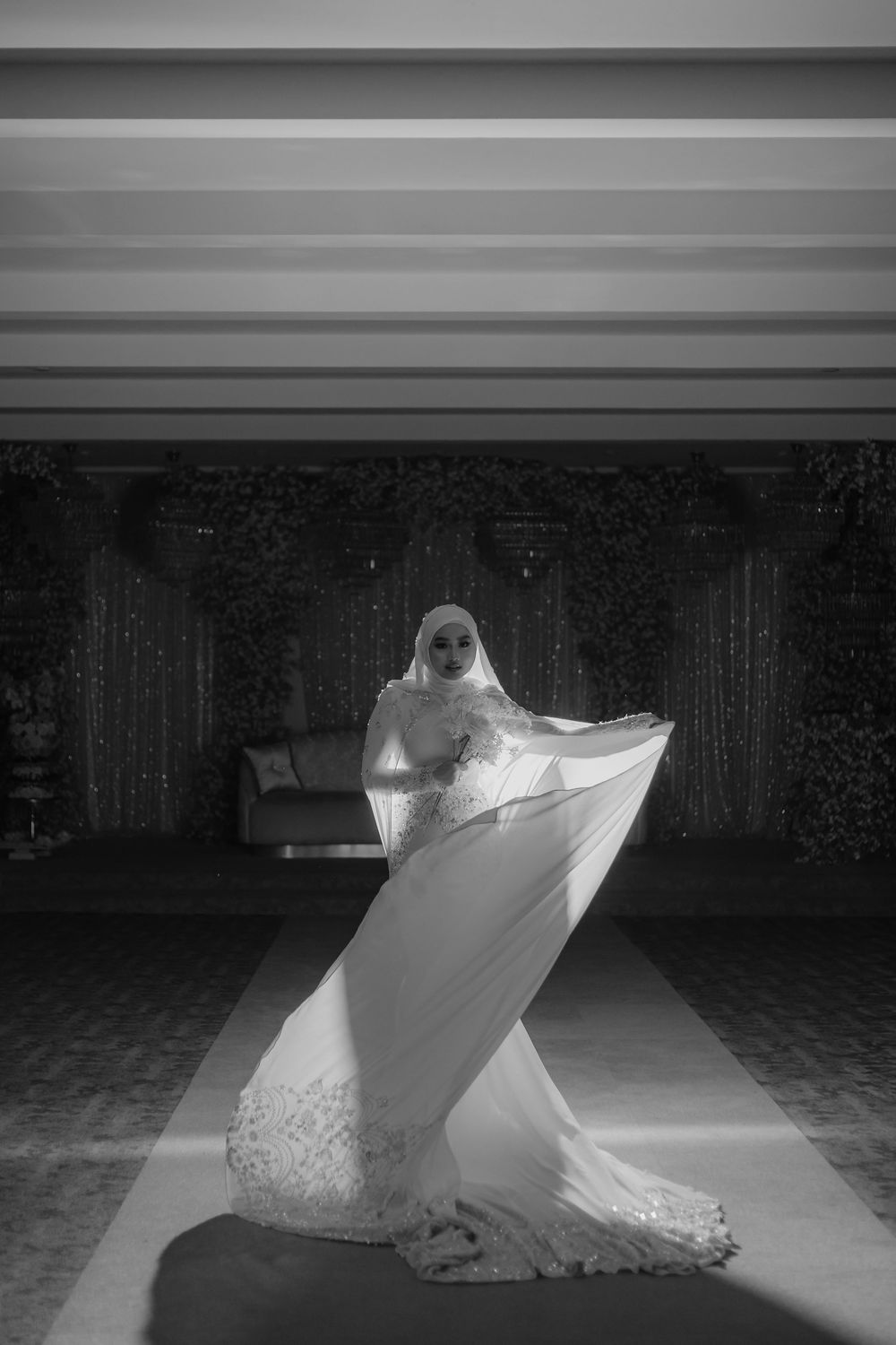 Black and white photo of a Muslim bride in hijab and lace wedding gown, elegantly posing with flowing veil.