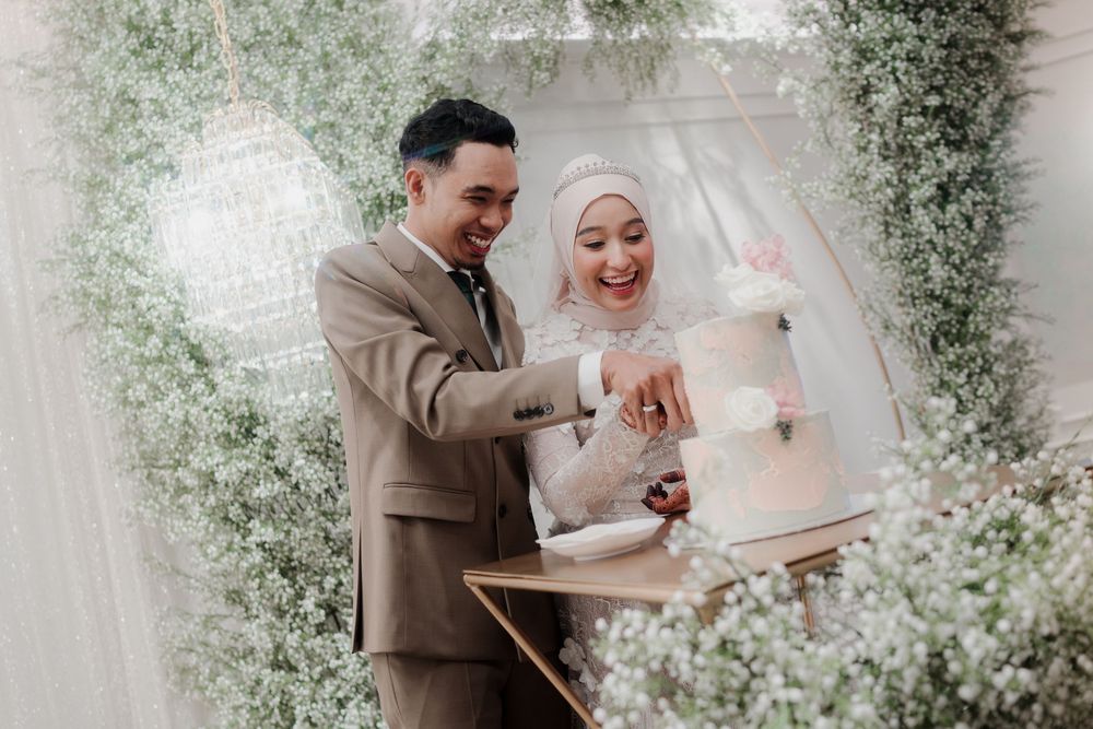 Muslim couple cutting wedding cake surrounded by lush greenery floral backdrop at elegant wedding ceremony.