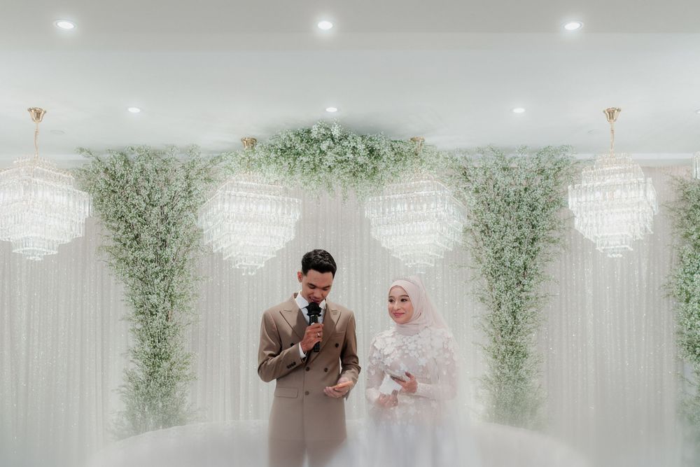 Muslim bride and groom stand before floral arch with crystal chandeliers at elegant white wedding ceremony.