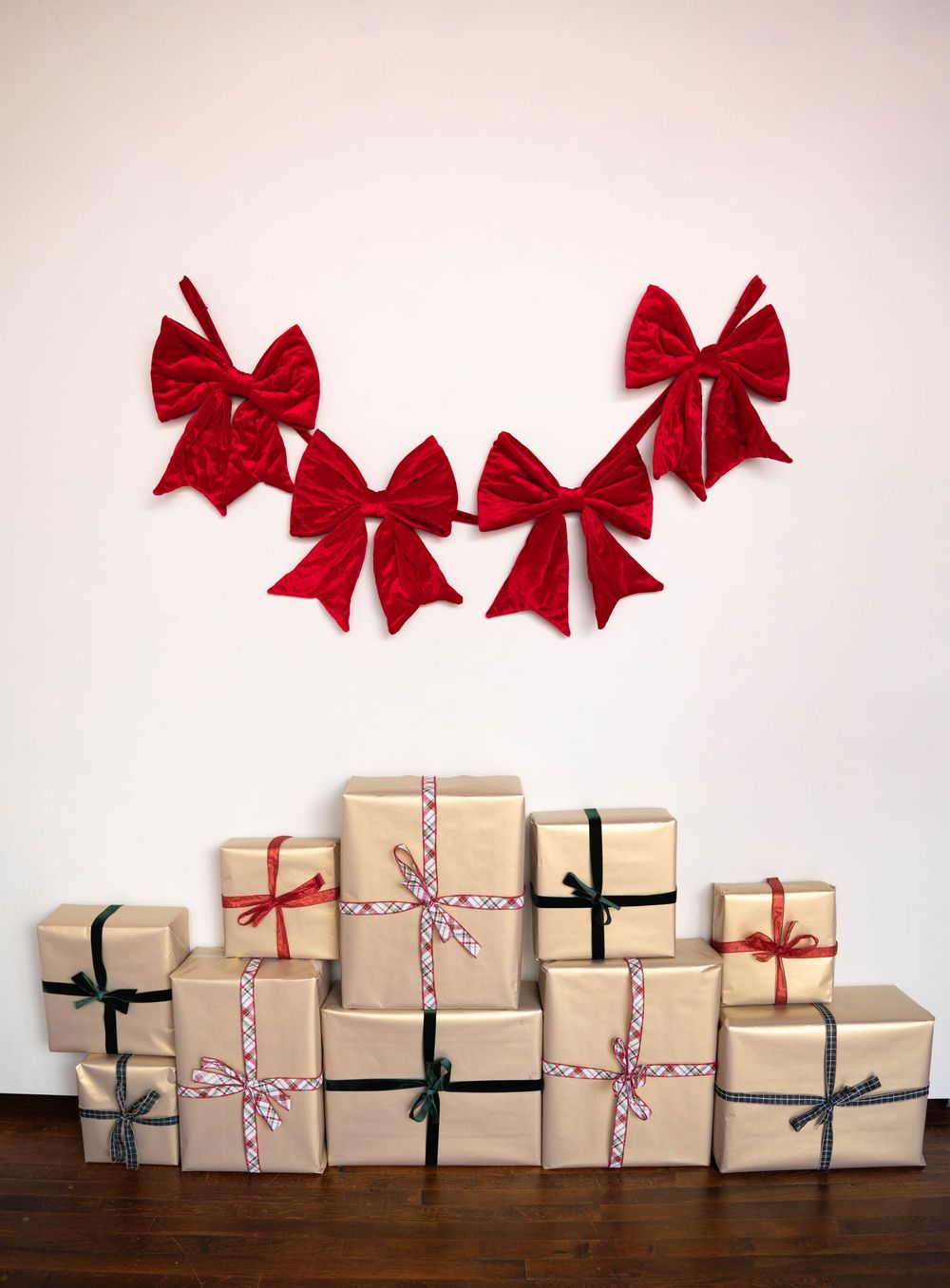 Stack of kraft paper wrapped Christmas gifts with colorful ribbons beneath a garland of red velvet bows on white wall.